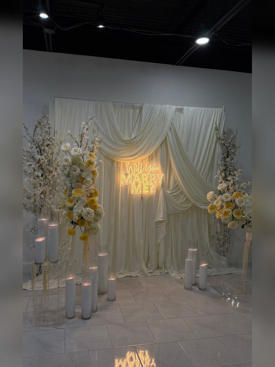 A draped backdrop with a glowing Will You Marry Me neon sign and pillar candles at The Hendrix