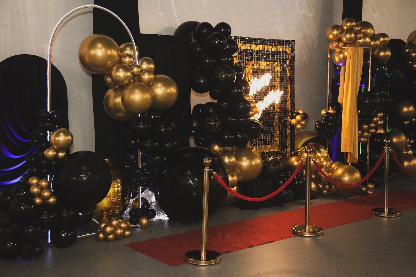 A red carpet with gold stanchions leading to a black and gold balloon and sequin backdrop at a birthday party at The Hendrix