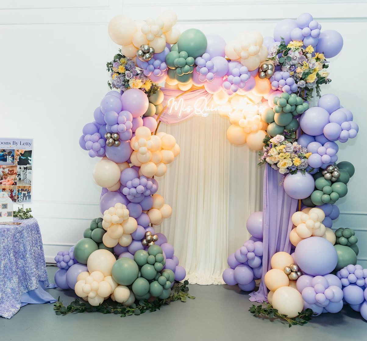 An organic balloon arch in sage, lilac and cream around a neon sign at The Hendrix