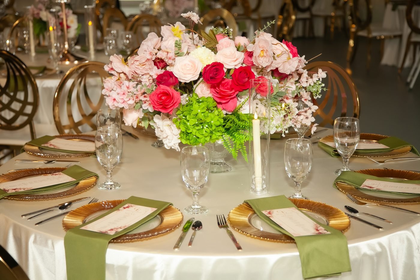 A round reception table with a pink and red rose centrepiece, gold charger plates, sage napkins and gold chairs at The Hendrix