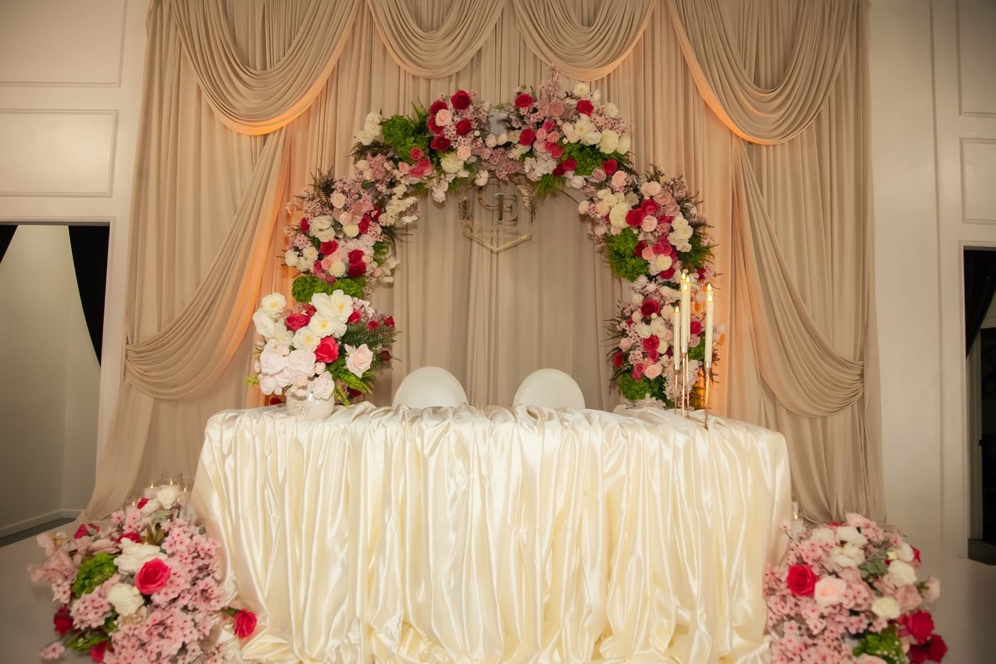 A sweetheart table under a pink and red floral arch with champagne drapes, set for a wedding reception in the Mansion at The Hendrix
